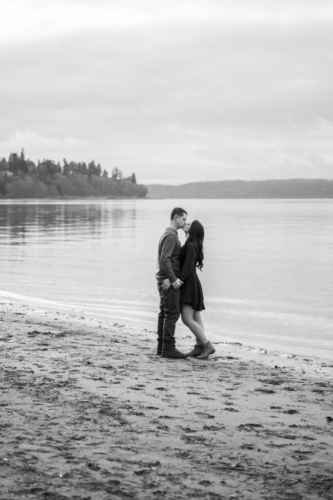 Engagement photography of a couple kissing on a beach at Picnic Point Park. The photograph is black and white.