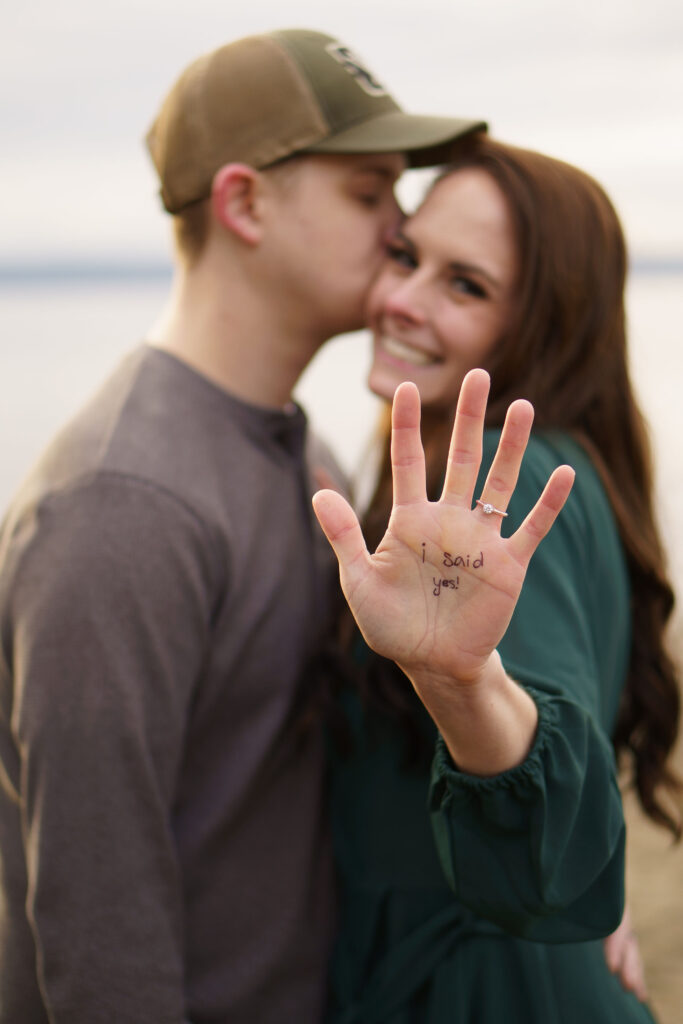 Engagement Photos of a couple at Picnic Point Park. A man is kissing his fiance's face, the fiance is looking at the camera with a big smile and holding out her palm to the camera. On her palm the words "I said yes!" are written, and it's the only thing in focus in the picture.