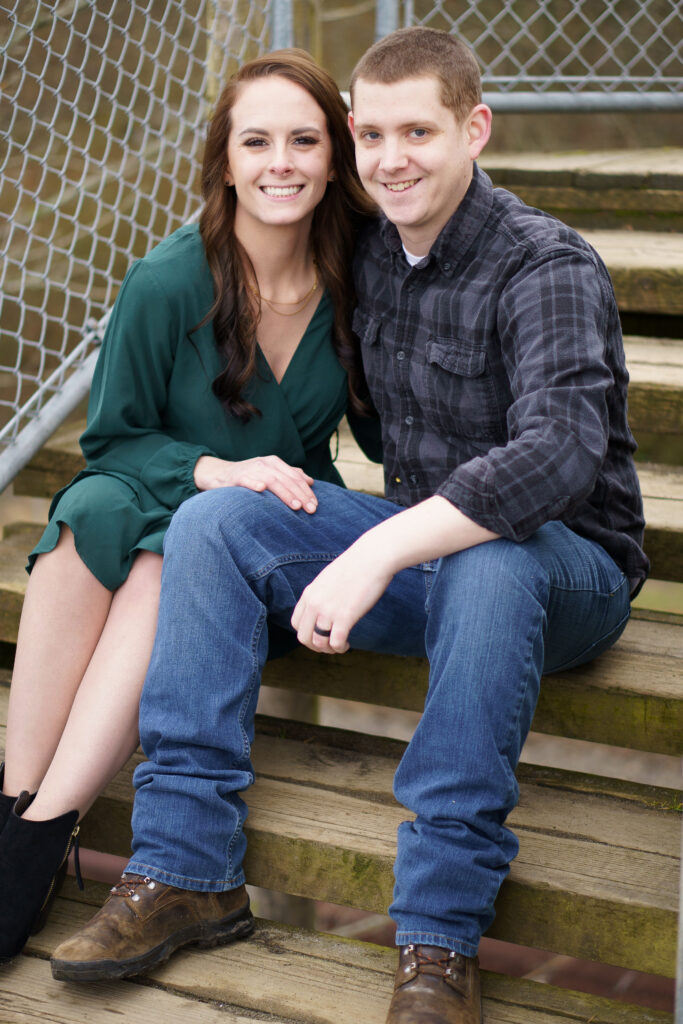 Engagement photos of a man and a woman sitting on wooden stairs, both smiling at the camera.