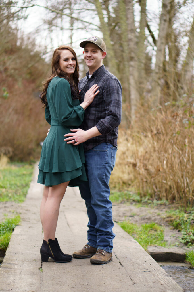 Engagement photos of a man and a woman standing on a wooden pathway surrounded by trees, standing closely, and smiling at the camera at Picnic Point Park.