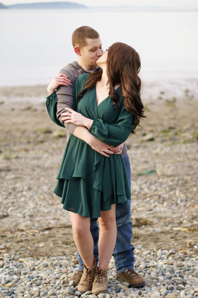 Engagement photo of a man and a woman kissing on a beach at Picnic Point Part while holding onto eachother.